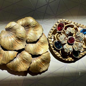 May include: Two vintage brooches. One is a cluster of bronze-colored leaf shapes. The other is a gold-toned brooch with a floral design, featuring red, blue, and clear gemstones. The brooches are likely from the mid-century era.