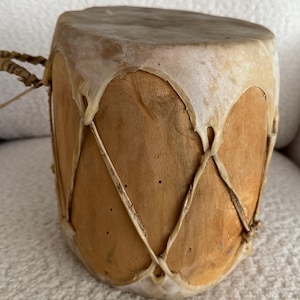 May include: A small, cylindrical drum with a natural wood body and a stretched, light-coloured hide head. The drum is bound with a crisscross pattern of natural cord, and a braided cord handle is visible.