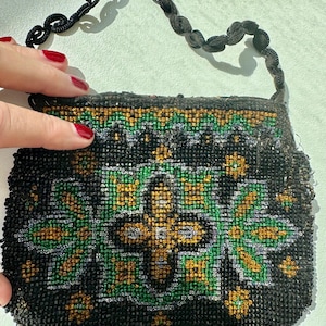 Pode incluir: Uma bolsa vintage com contas com fundo preto e um padrão floral em verde e dourado. A bolsa tem uma alça de contas pretas. A bolsa é segurada por uma mão com esmalte vermelho.