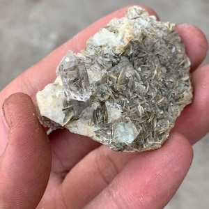 Puede incluir: Un espécimen mineral en bruto con un grupo de cristales. La piedra presenta una mezcla de colores claros, blancos y grises. Los cristales están parcialmente incrustados en una matriz de minerales en capas y escamosos. La textura general es áspera y natural.