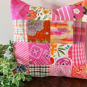 May include: A pink and white patchwork pillow with a variety of floral and geometric patterns. The pillow is made from different fabrics and is quilted.