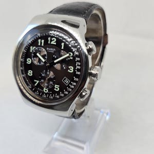 May include: A Swatch chronograph watch with a silver-tone metal case, a black dial featuring white Arabic numerals, three sub-dials, a date window at the four o'clock position, and a black leather strap with a textured finish and silver buckle.
