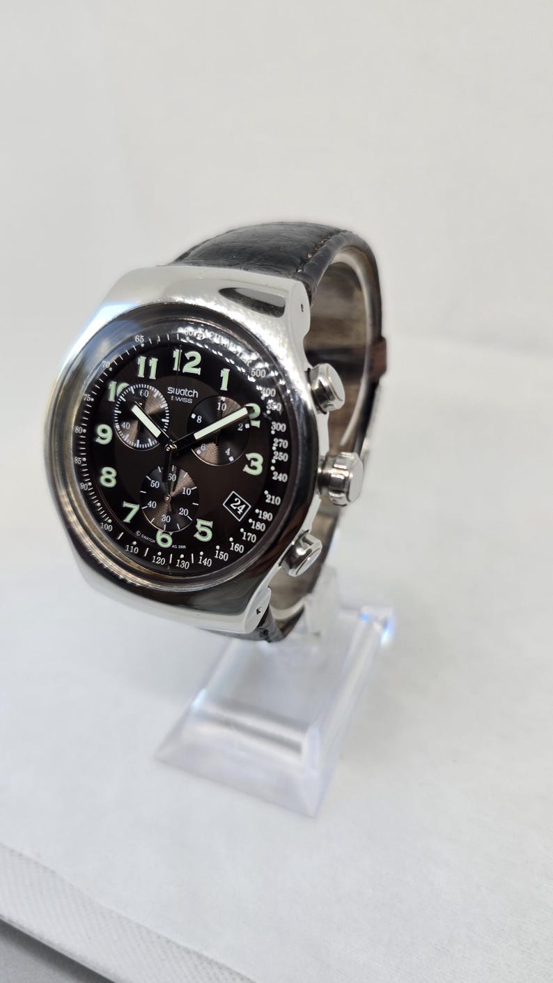 May include: A Swatch chronograph watch with a silver-tone metal case, a black dial featuring white Arabic numerals, three sub-dials, a date window at the four o'clock position, and a black leather strap with a textured finish and silver buckle.