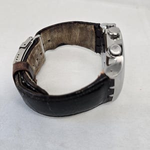 May include: A wristwatch featuring a polished silver-tone metal case and a dark brown leather strap with a visible metal deployment clasp. The interior of the leather strap shows signs of wear with a light tan, textured surface.