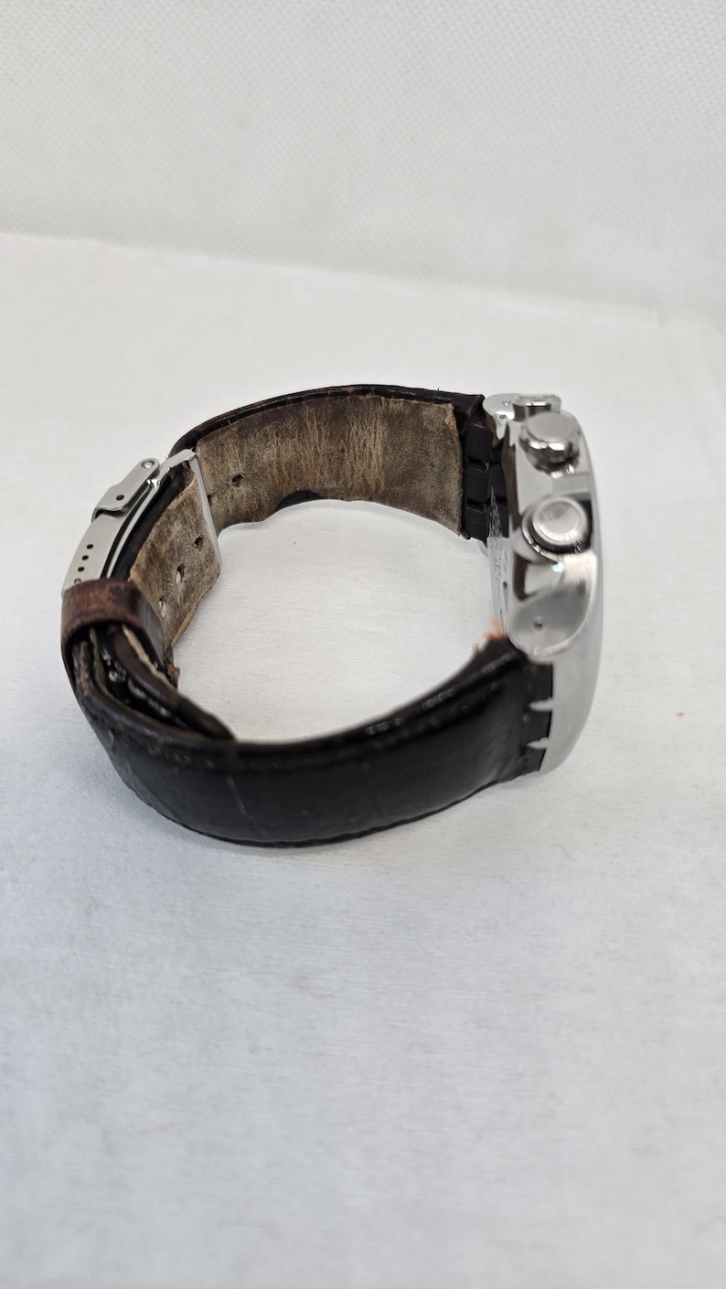 May include: A wristwatch featuring a polished silver-tone metal case and a dark brown leather strap with a visible metal deployment clasp. The interior of the leather strap shows signs of wear with a light tan, textured surface.