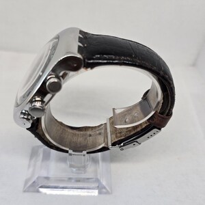 May include: A wristwatch with a silver-toned metal case and a black textured leather strap. The side of the watch features two circular pushers and a central crown. The strap includes a silver-toned metal deployment clasp mechanism.