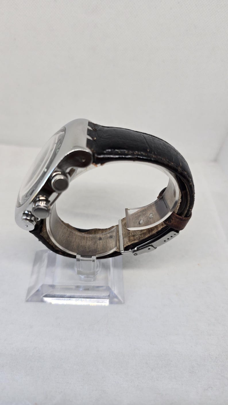 May include: A wristwatch with a silver-toned metal case and a black textured leather strap. The side of the watch features two circular pushers and a central crown. The strap includes a silver-toned metal deployment clasp mechanism.