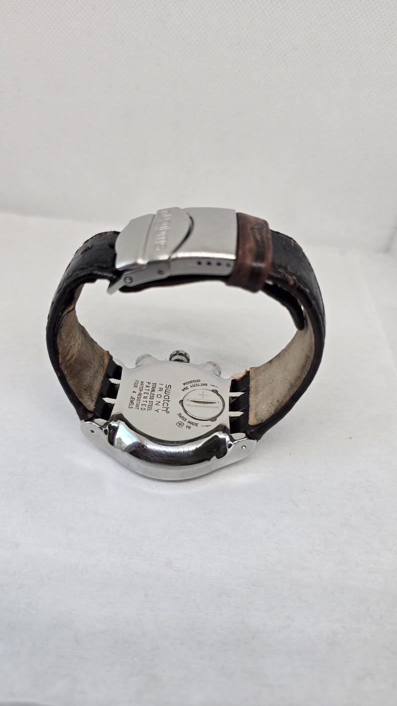 May include: A Swatch Irony stainless steel watch with a dark brown leather strap and a silver-tone metal deployment clasp. The back of the watch case features engraved text including Swatch Irony, stainless steel, patented, water-resistant, and four jewels.