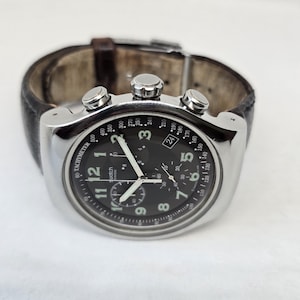 May include: A silver-tone chronograph wristwatch with a black dial, white Arabic numerals, and three sub-dials. The watch features a tachymeter scale on the outer rim, luminous hands, and a dark brown leather strap with a metal buckle closure.