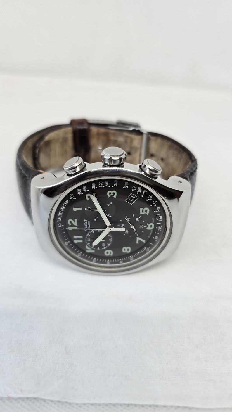 May include: A silver-tone chronograph wristwatch with a black dial, white Arabic numerals, and three sub-dials. The watch features a tachymeter scale on the outer rim, luminous hands, and a dark brown leather strap with a metal buckle closure.