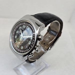 May include: A silver-tone chronograph wristwatch featuring a black dial with white Arabic numerals, three sub-dials, and a date window at the four o'clock position. The watch includes a tachymeter scale on the bezel and a dark brown leather strap.