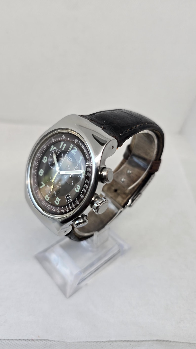 May include: A silver-tone chronograph wristwatch featuring a black dial with white Arabic numerals, three sub-dials, and a date window at the four o'clock position. The watch includes a tachymeter scale on the bezel and a dark brown leather strap.