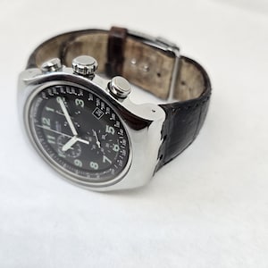 May include: A silver-tone chronograph wristwatch with a black dial, white Arabic numerals, and three sub-dials. The watch features a date window at the four o'clock position, a tachymeter scale on the outer bezel, and a dark brown leather strap.