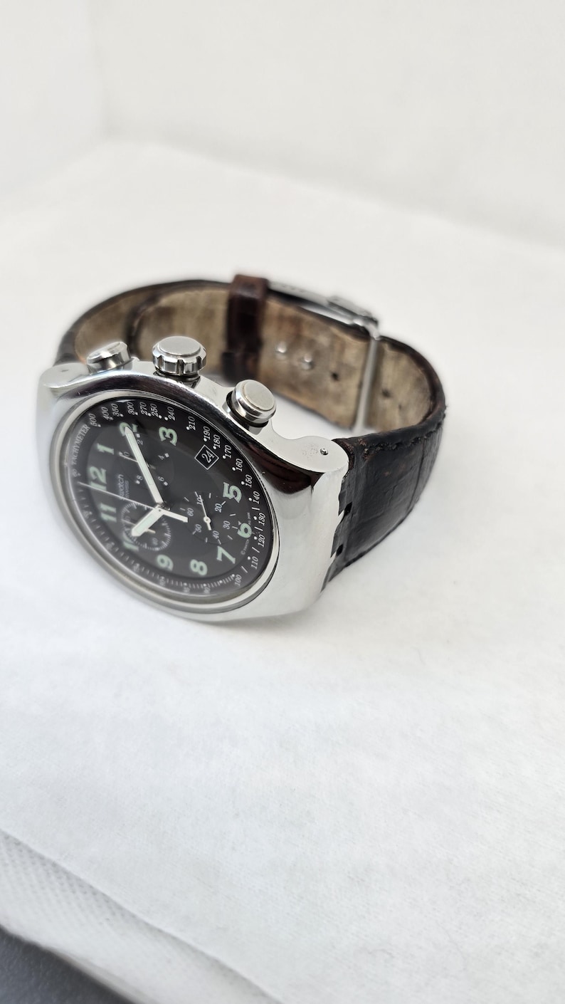 May include: A silver-tone chronograph wristwatch with a black dial, white Arabic numerals, and three sub-dials. The watch features a date window at the four o'clock position, a tachymeter scale on the outer bezel, and a dark brown leather strap.