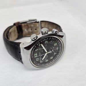 May include: A silver-tone chronograph wristwatch with a black dial, white Arabic numerals, and three sub-dials. The watch features a tachymeter scale on the outer bezel, a date window, and a dark brown leather strap with a metal deployment clasp.