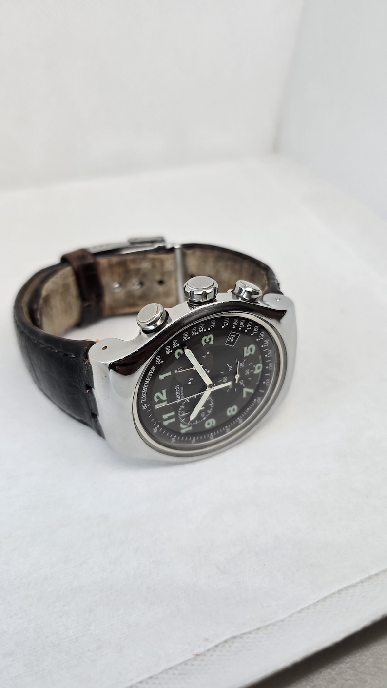 May include: A silver-tone chronograph wristwatch with a black dial, white Arabic numerals, and three sub-dials. The watch features a tachymeter scale on the outer bezel, a date window, and a dark brown leather strap with a metal deployment clasp.