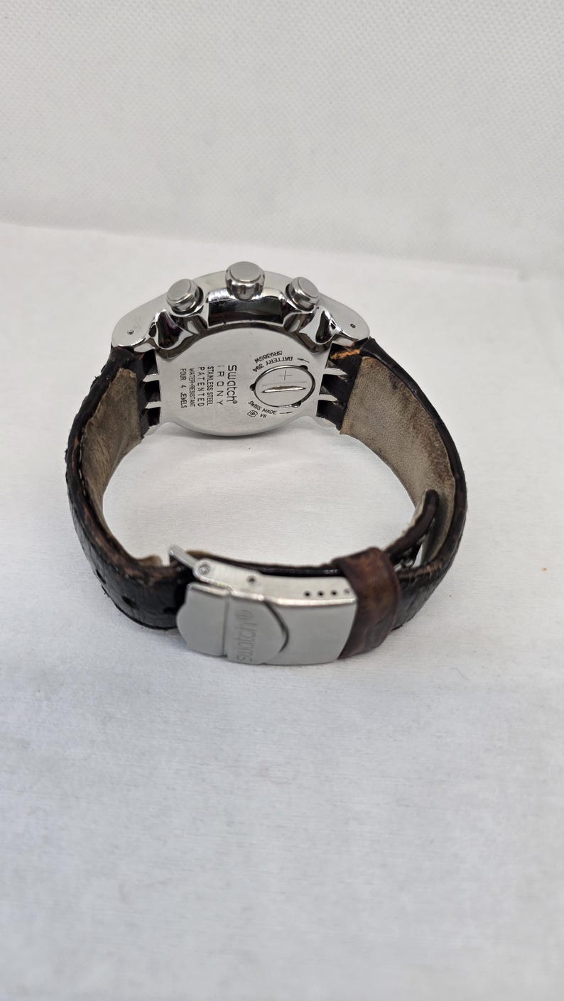 May include: A Swatch Irony stainless steel watch with a brown textured leather strap and a silver-tone metal deployment clasp. The back of the watch case features the text Swatch Irony, Stainless Steel, Patented, Water-Resistant, Four 4 Jewels.