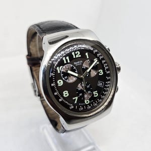 May include: A silver-tone chronograph wristwatch with a black dial, featuring white Arabic numerals, three sub-dials, a date window at the three o'clock position, and a black leather strap. The dial displays the Swatch brand name and tachymeter scale.