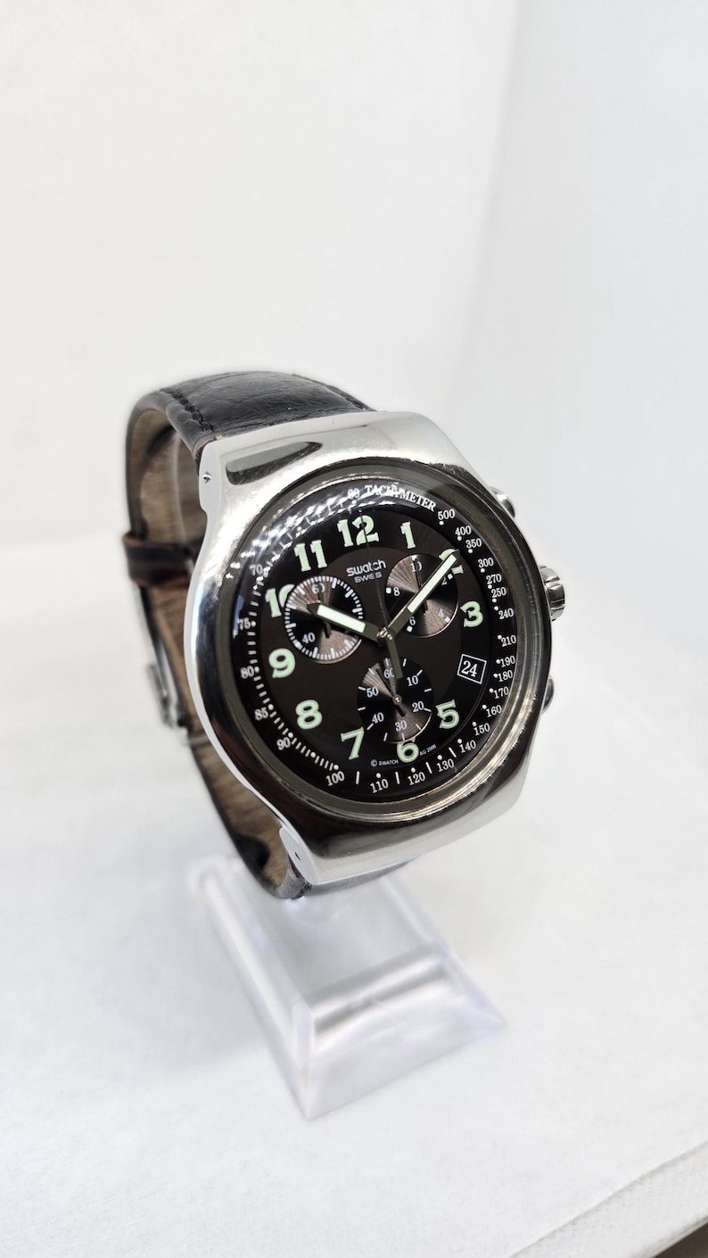 May include: A silver-tone chronograph wristwatch with a black dial, featuring white Arabic numerals, three sub-dials, a date window at the three o'clock position, and a black leather strap. The dial displays the Swatch brand name and tachymeter scale.