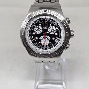 May include: A silver-tone stainless steel Swatch chronograph wristwatch with a black dial, featuring a world time bezel with city names, three sub-dials, a red central second hand, and a metal link bracelet with a deployment clasp closure.