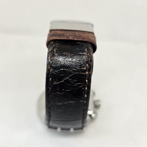 May include: A wristwatch featuring a weathered black textured strap with visible cracks and a brown leather accent near the silver-toned metal buckle. The watch case is partially visible behind the strap, showing small silver side buttons.