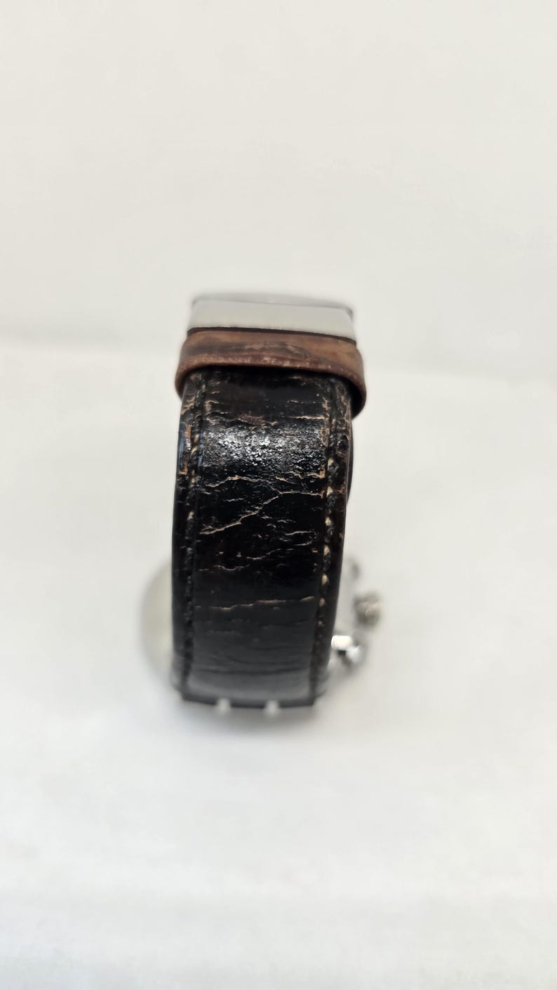 May include: A wristwatch featuring a weathered black textured strap with visible cracks and a brown leather accent near the silver-toned metal buckle. The watch case is partially visible behind the strap, showing small silver side buttons.