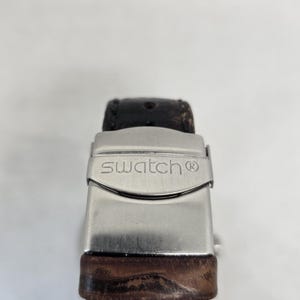 May include: A dark brown textured leather watch strap featuring a silver-toned metal deployment clasp. The clasp is engraved with the word swatch and a registered trademark symbol. The leather strap shows visible signs of wear and grain texture.