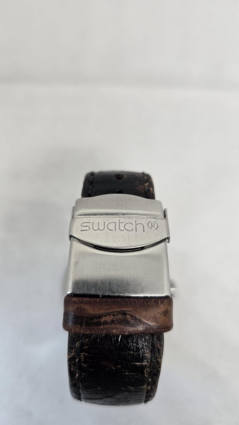 May include: A dark brown textured leather watch strap featuring a silver-toned metal deployment clasp. The clasp is engraved with the word swatch and a registered trademark symbol. The leather strap shows visible signs of wear and grain texture.
