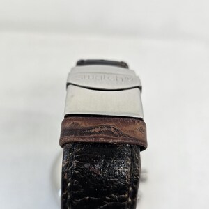 May include: A wristwatch with a dark brown, textured leather strap and a silver-toned metal clasp. The clasp features the word swatch engraved on its surface. The strap shows visible signs of wear with a cracked texture throughout the leather.