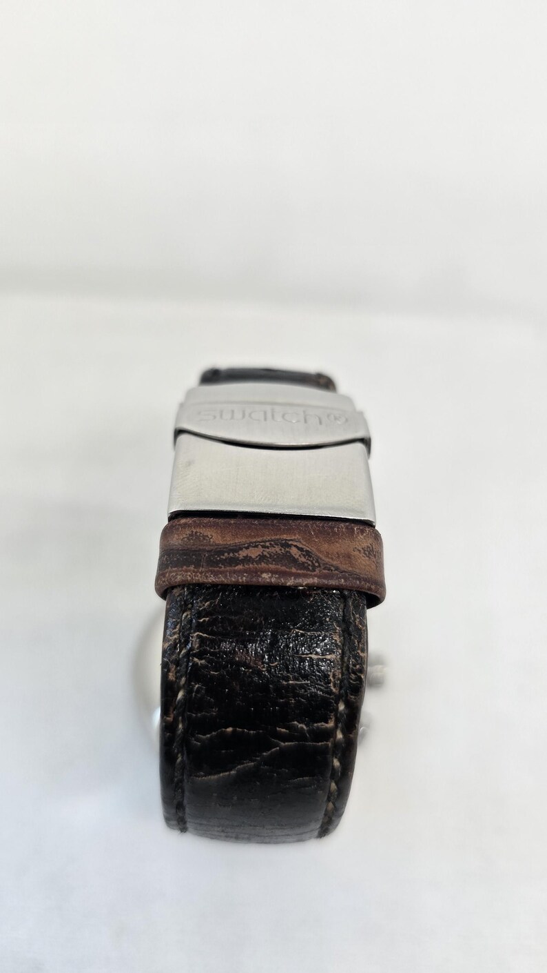 May include: A wristwatch with a dark brown, textured leather strap and a silver-toned metal clasp. The clasp features the word swatch engraved on its surface. The strap shows visible signs of wear with a cracked texture throughout the leather.