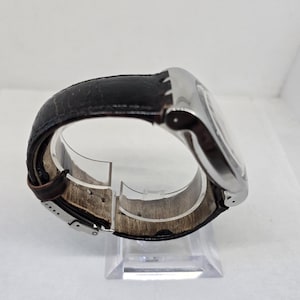 May include: A wristwatch featuring a polished silver-tone metal case and a dark brown leather strap with a visible texture. The strap includes a silver-tone metal buckle clasp mechanism, showing the interior lining and the side profile of the watch.