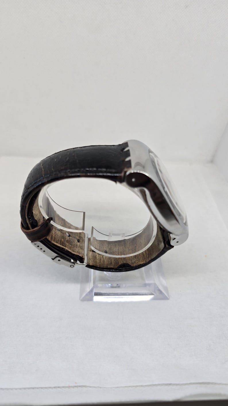 May include: A wristwatch featuring a polished silver-tone metal case and a dark brown leather strap with a visible texture. The strap includes a silver-tone metal buckle clasp mechanism, showing the interior lining and the side profile of the watch.
