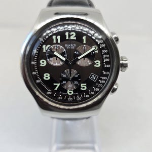 May include: A Swatch chronograph watch with a silver-tone metal case and a black leather strap. The black dial features white Arabic numerals, three sub-dials, a date window at the four o'clock position, and a tachymeter scale on the outer bezel.
