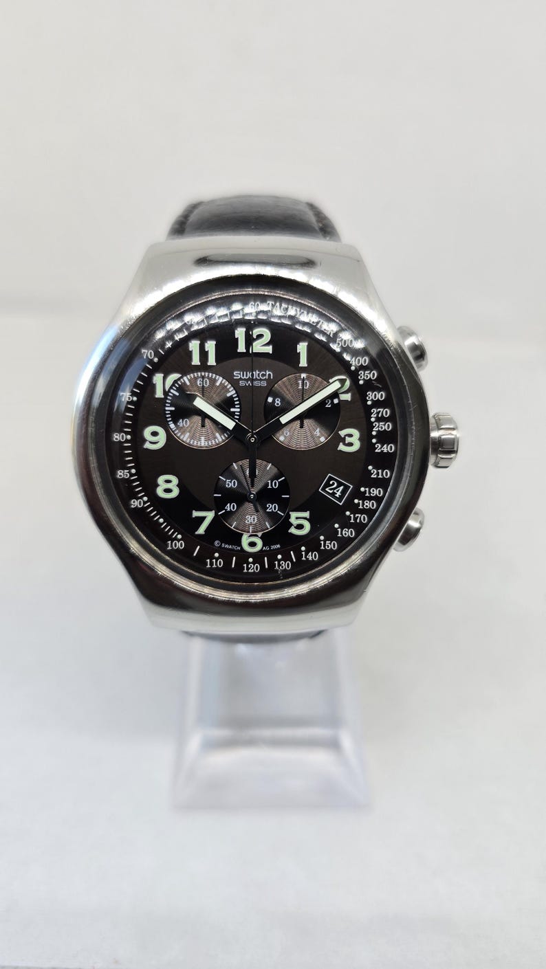 May include: A Swatch chronograph watch with a silver-tone metal case and a black leather strap. The black dial features white Arabic numerals, three sub-dials, a date window at the four o'clock position, and a tachymeter scale on the outer bezel.
