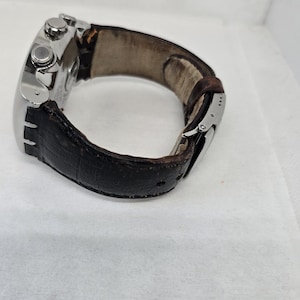 May include: A wristwatch featuring a silver-toned metal case and a dark brown textured leather strap. The watch includes a side crown and two pushers, with a metal deployment clasp visible on the strap. The interior of the strap shows signs of wear.
