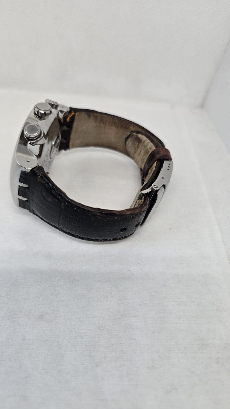 May include: A wristwatch featuring a silver-toned metal case and a dark brown textured leather strap. The watch includes a side crown and two pushers, with a metal deployment clasp visible on the strap. The interior of the strap shows signs of wear.
