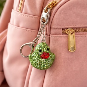 May include: A green rhinestone duck keychain with a red mouth and black eyes hangs from a silver chain attached to a pink backpack. The keychain is a decorative accessory.