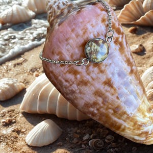 May include: A silver chain bracelet with a round, patterned charm is draped over a large, pink and brown seashell. The bracelet is resting on a sandy beach with other seashells and the ocean in the background. The charm has a unique, speckled design.
