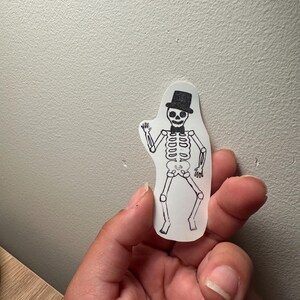 Op de afbeelding: Een doorzichtige sticker met een zwart-witte illustratie van een skelet met een hoge hoed en een vlinderdas. Het skelet staat in een danshouding, met één arm omhoog.
