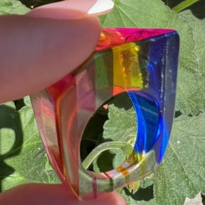 Peut inclure: Une bague épaisse et translucide présentant un design dégradé aux couleurs vives. L'anneau affiche un spectre de teintes incluant le bleu profond, le jaune vif, le vert et le magenta, formant une silhouette géométrique aux bords arrondis.