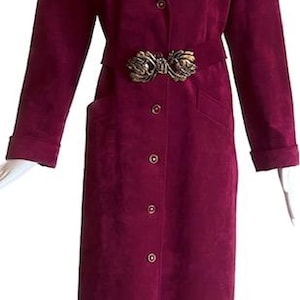 May include: A long, burgundy suede coat with a notched collar, button closure, and a decorative gold-tone belt buckle. The coat features long sleeves, pockets, and a tailored fit, creating a sophisticated silhouette. This vintage coat is a stylish outerwear option.