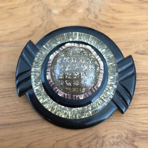 May include: A black and white plastic brooch with a round, glittery, gold-toned center.