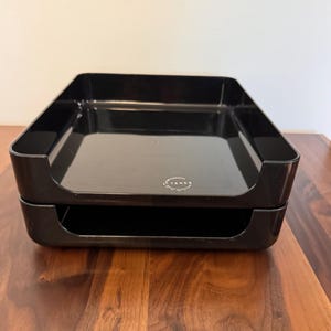 May include: Two-tier black plastic desk organizer. The organizer has a glossy finish and a rectangular shape with rounded corners. The brand name "TENEX" is printed in white on the front of the top tray. The trays are stacked on top of each other.