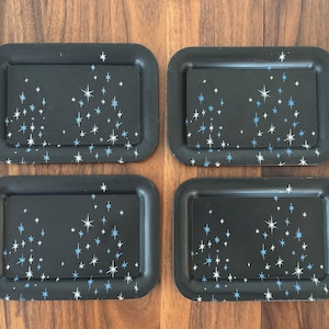 May include: Set of four black rectangular trays with rounded corners. Each tray features a pattern of white and blue starbursts against a black background. The trays are arranged on a wooden surface.