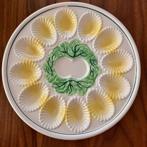May include: A round white ceramic serving platter featuring twelve shell-shaped indentations arranged in a circle. Each indentation has a pale yellow center. The center of the plate features a raised green leaf design with dark green line details.