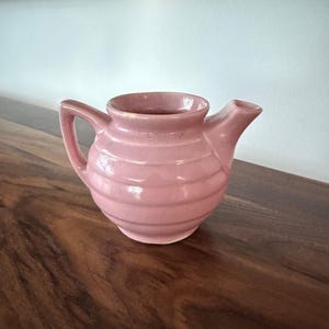 May include: A pink ceramic teapot with a handle and spout. The teapot has a ribbed design around the body and sits on a wooden surface. The teapot is a soft, muted pink color.