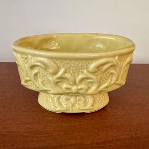 May include: A pale yellow ceramic bowl with a pedestal base. The bowl features embossed floral and scrollwork designs around the sides and base. The bowl is approximately 4 inches in diameter and 3 inches tall.