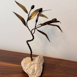 May include: A decorative metal sculpture of an olive branch with brass-toned leaves and dark spherical olives. The branch features a twisted metal stem that is securely anchored into a rough, light-colored natural stone base.