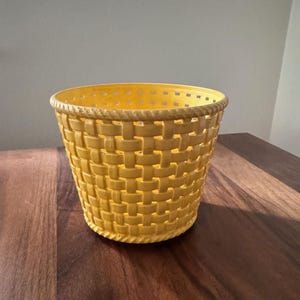 May include: A yellow woven basket with a cylindrical shape. The basket has a textured, basket-weave pattern and a decorative rope-like trim around the top. The basket is sitting on a dark wooden surface.