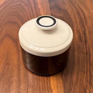 May include: A round, lidded container with a white top and a dark brown base. The lid has a black and white circular handle. The container is sitting on a wooden surface. This item is suitable for storing small items.